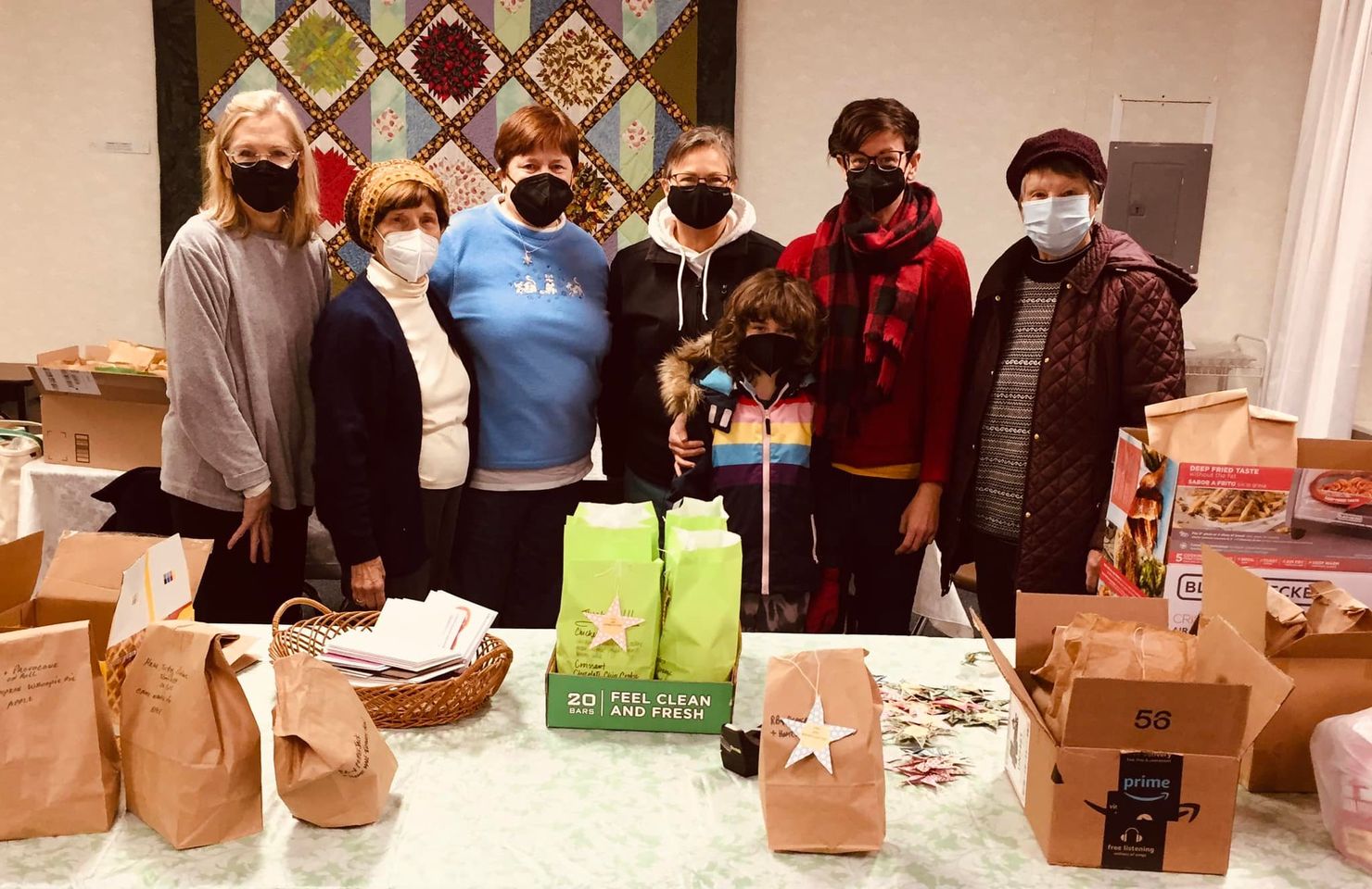 Photo of six church members and a child standing behind a table that holds bagged lunches and a box of greeting cards.